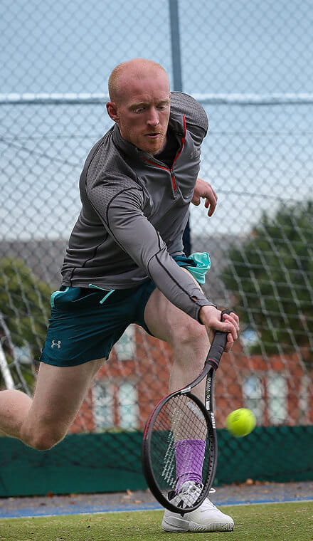 A colour photo shows a Royal Navy tennis player on an outdoor court. He swings his racket to hit a low spinning ball.
