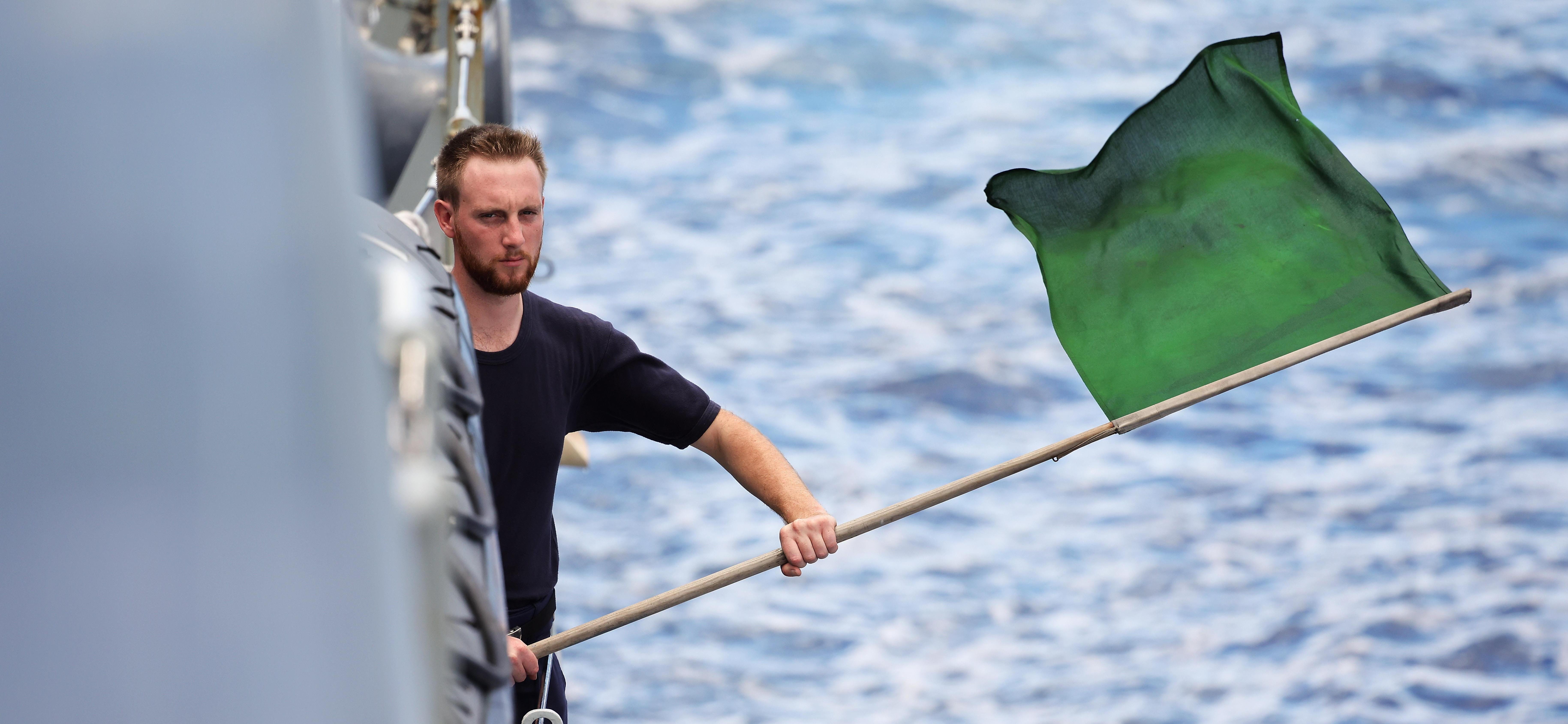 A Royal Navy sailor waves a green flag. Boats crews onboard HMS Tamar prepare to launch a seaboat to conduct a search and rescue exercise.